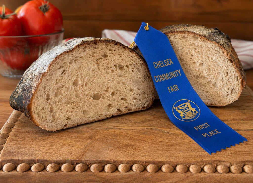 A beautiful sourdough loaf with an excellent crumb on a wood cutting board with a blue ribbon from the Chelsea Community Fair