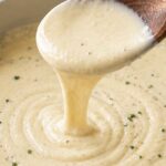 A wooden spoon lifting creamy Alfredo sauce from a skillet