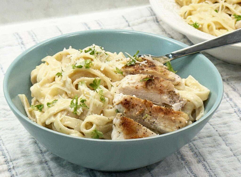 Chicken alfredo with sliced seared chicken over creamy fettucinne in a teal blue bowl, topped with grated Parmesan and parsley