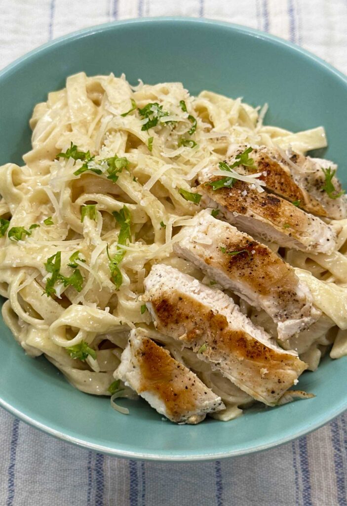 Chicken alfredo with sliced seared chicken over creamy fettucinne in a teal blue bowl, topped with grated Parmesan and parsley