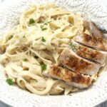 Chicken alfredo with sliced seared chicken over creamy fettucinne in a white bowl, topped with grated Parmesan and parsley