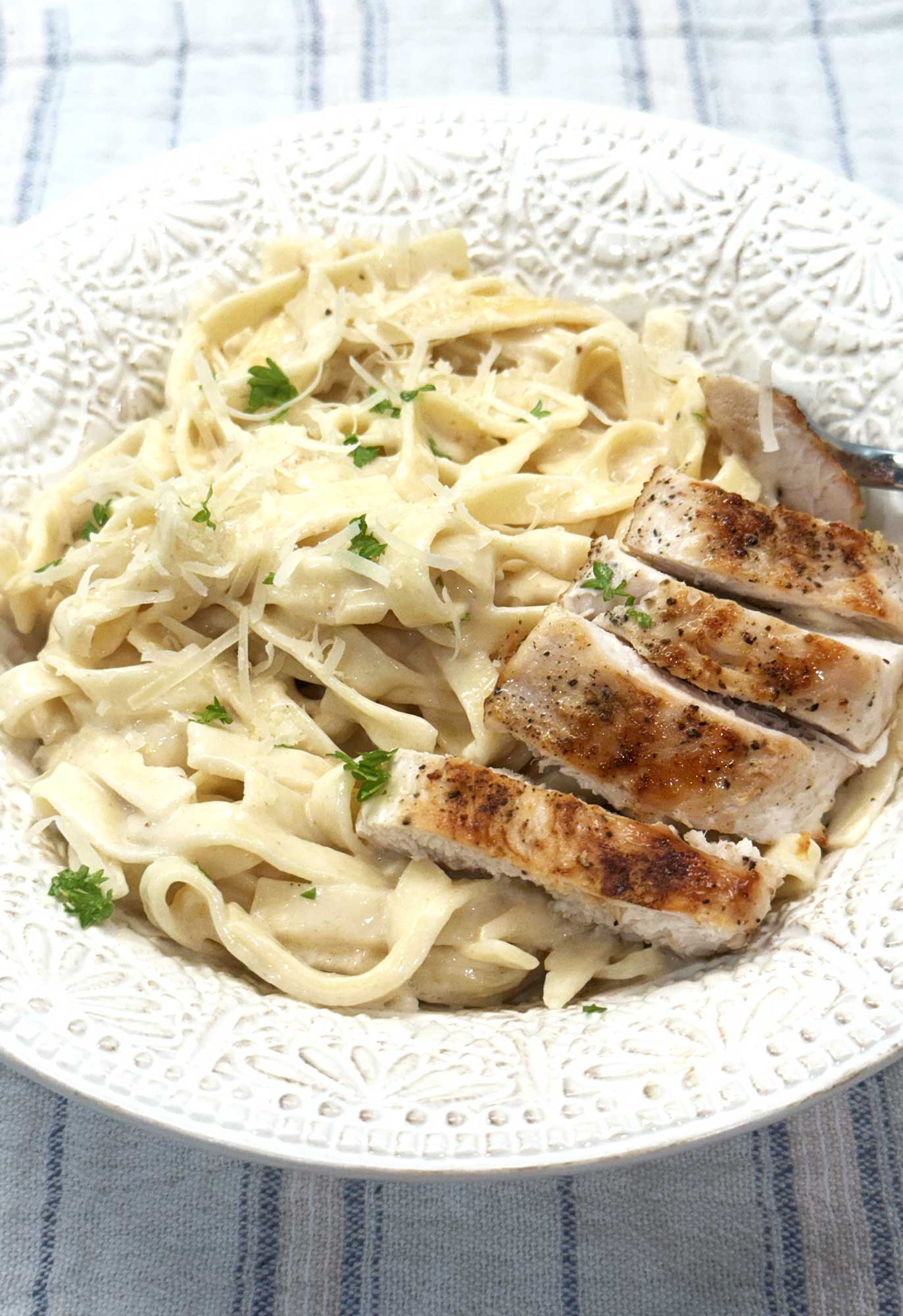 Chicken alfredo with sliced seared chicken over creamy fettucinne in a white bowl, topped with grated Parmesan and parsley