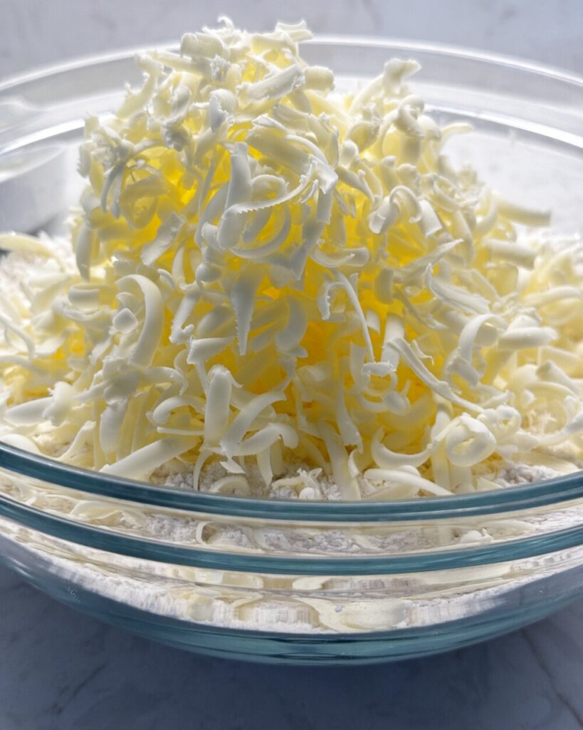 Grated frozen butter shreds tossed into a glass bowl of flour for sourdough discard biscuits