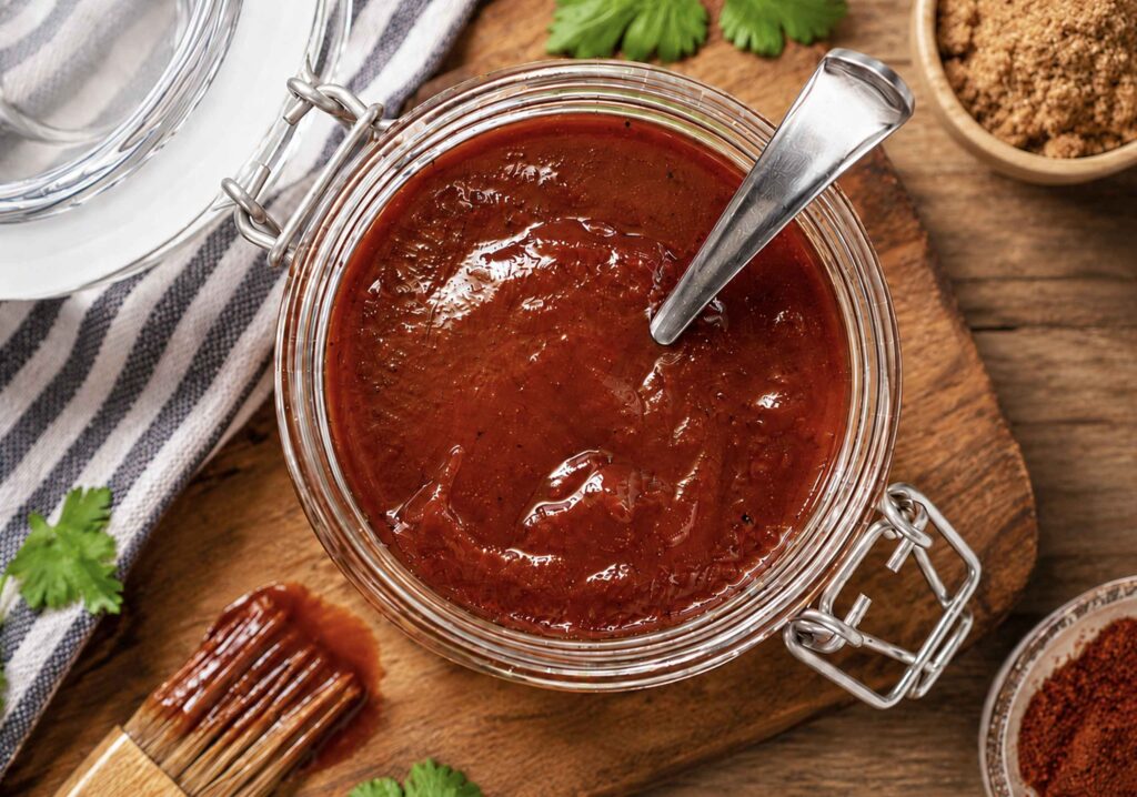 Homemade bbq sauce in a jar beside bowls of ingredients and a brush