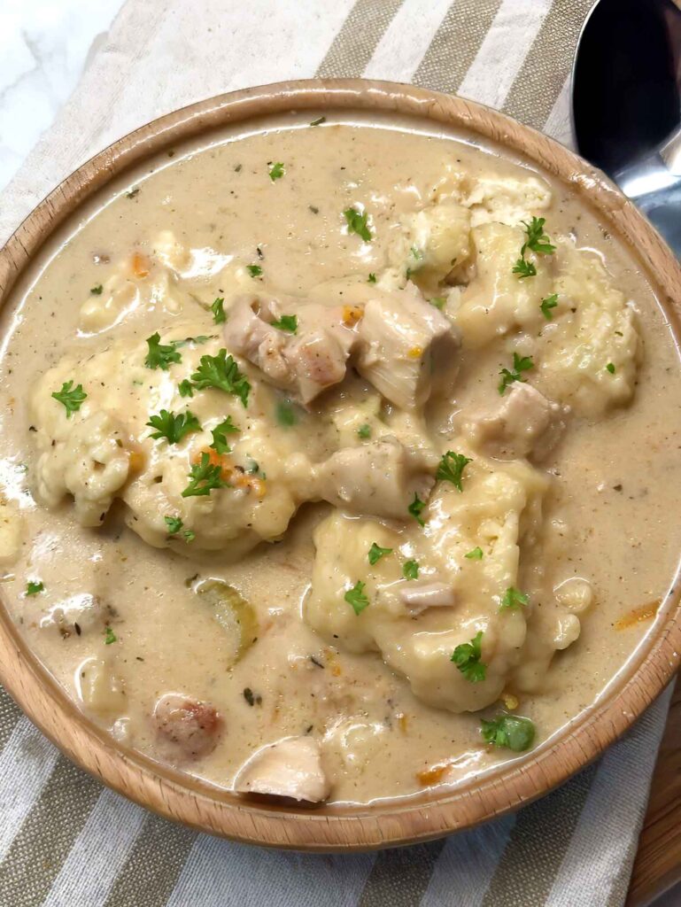 A wooden bowl of chicken and dumplings with soft dumplings, tender chicken, and fresh parsley in a creamy broth