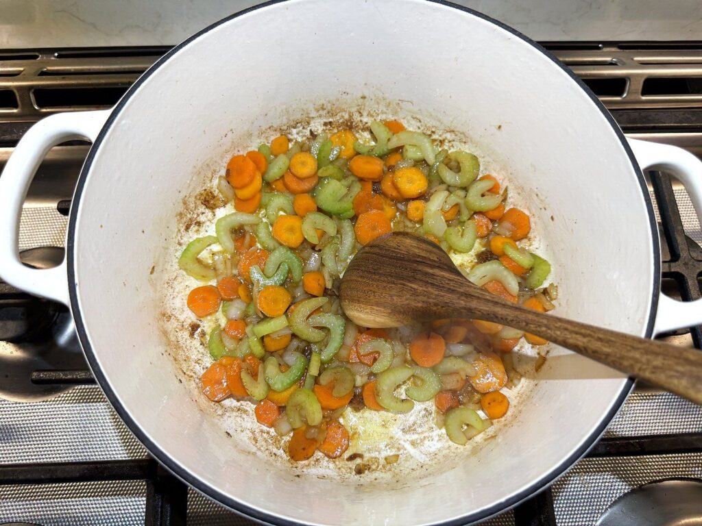 Carrots, celery, and onion cooking with a wooden spoon in a white pot to build the chicken stew