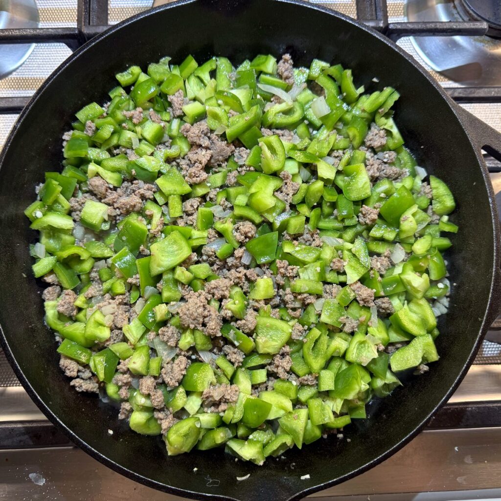 Chopped green bell peppers added to browned beef in a cast iron skillet.