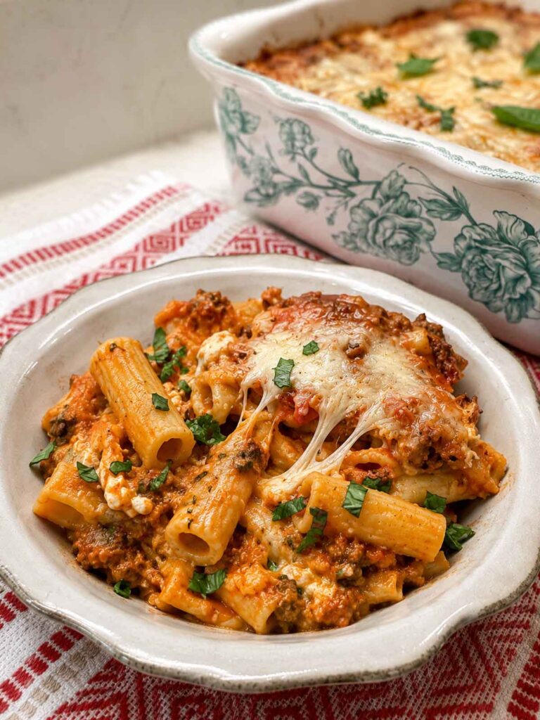 A bowl of baked ziti with a cheese pull, the baking dish in the background