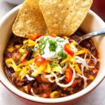 Bowl of taco soup topped with tortilla chips, shredded cheese, sour cream, and red pepper
