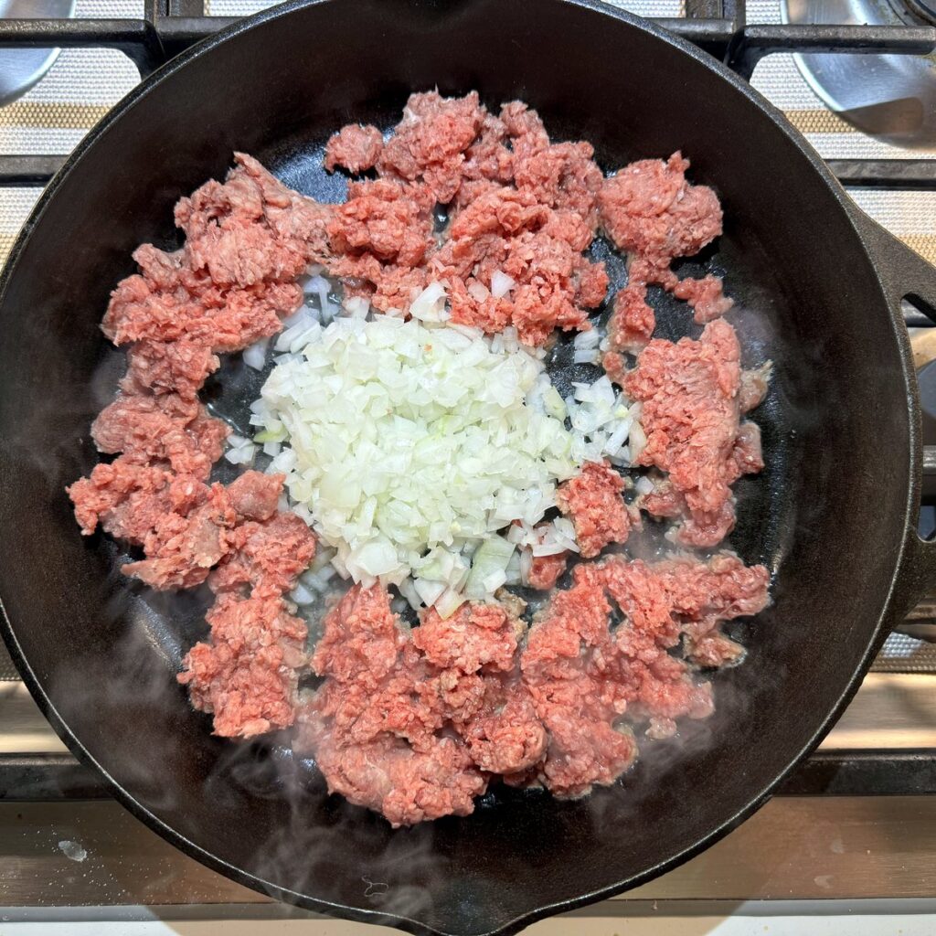Ground beef and diced onion browning together in a cast iron skillet.