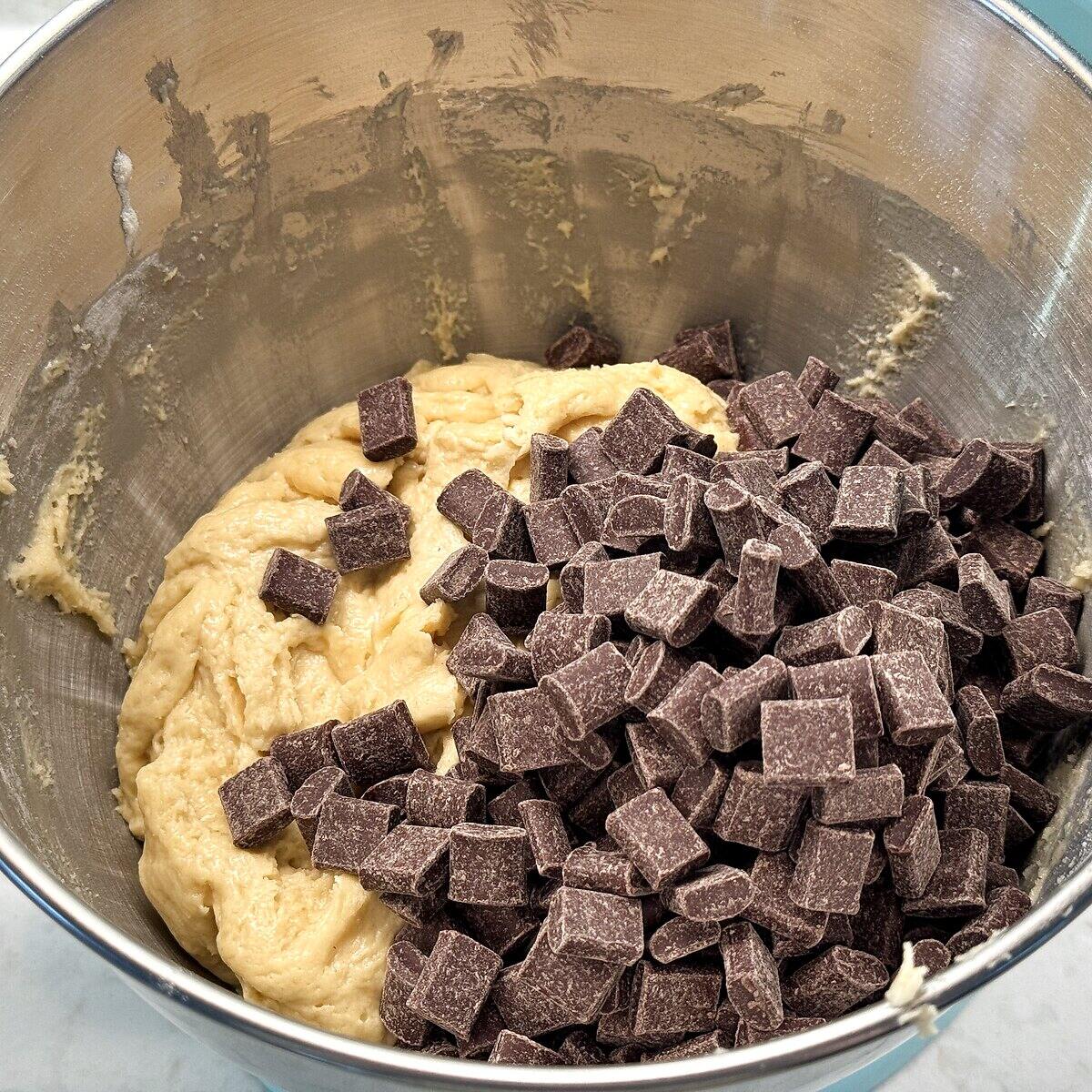 A big pile of chocolate chunks on top of sourdough cookie dough in a stand mixer bowl.