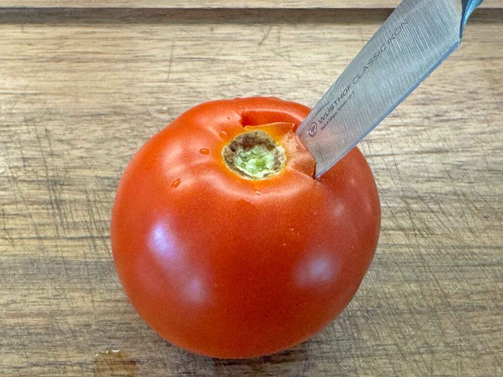 A fresh tomato being cored with a knife on a cutting board
