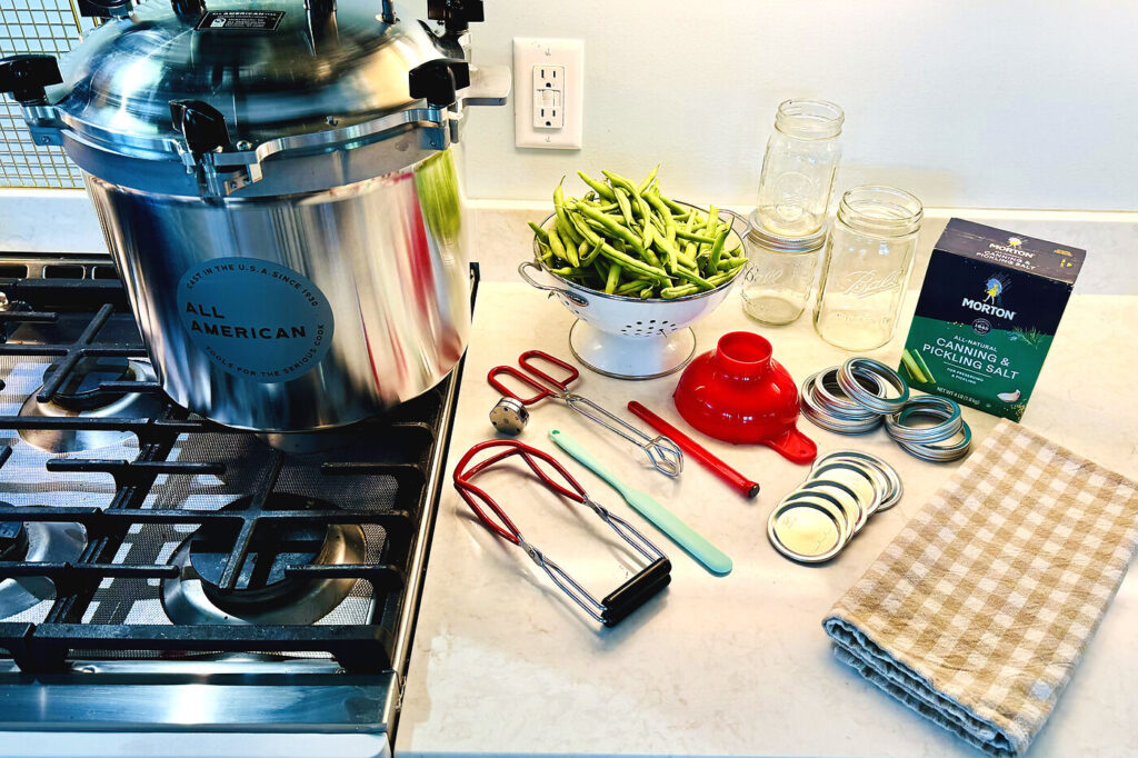 An All American pressure canner on the stove with jars, lids, jar lifter, funnel, and fresh green beans laid out on the counter