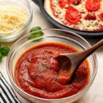A glass bowl of thick, smooth homemade pizza sauce with a wooden spoon resting in it