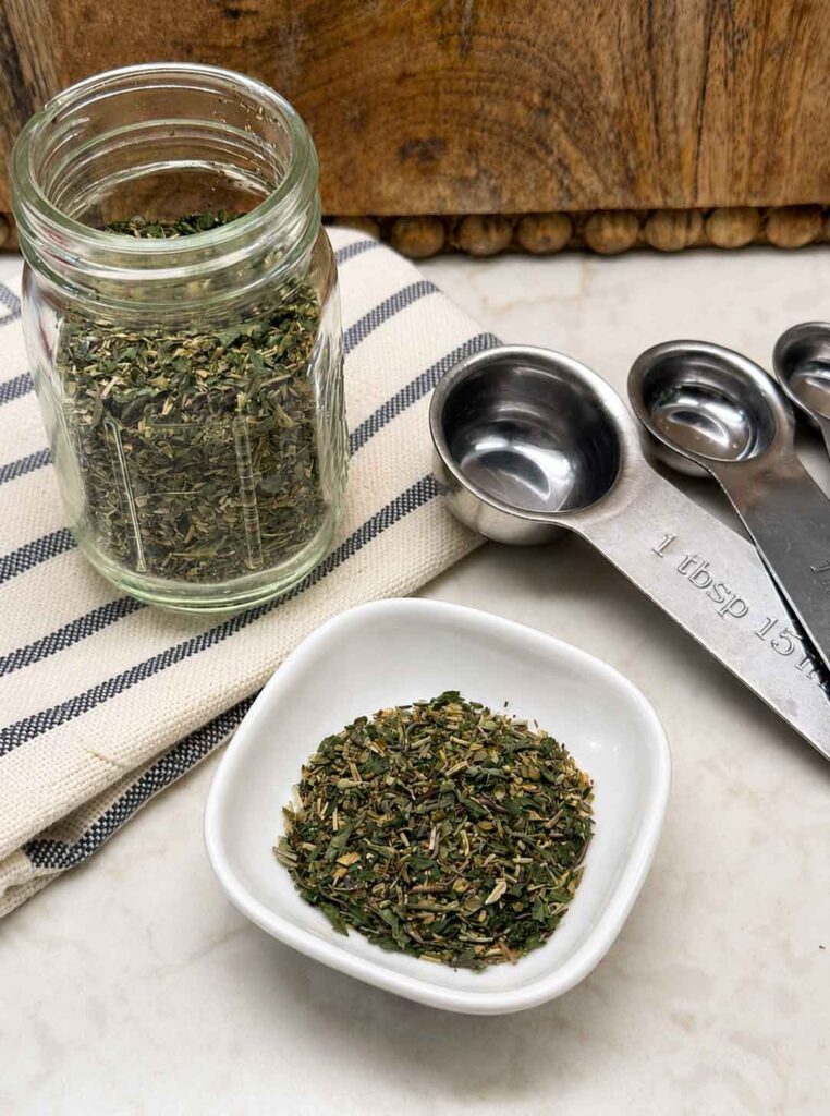 A jar of homemade Italian seasoning with a small dish of the blend and measuring spoons.