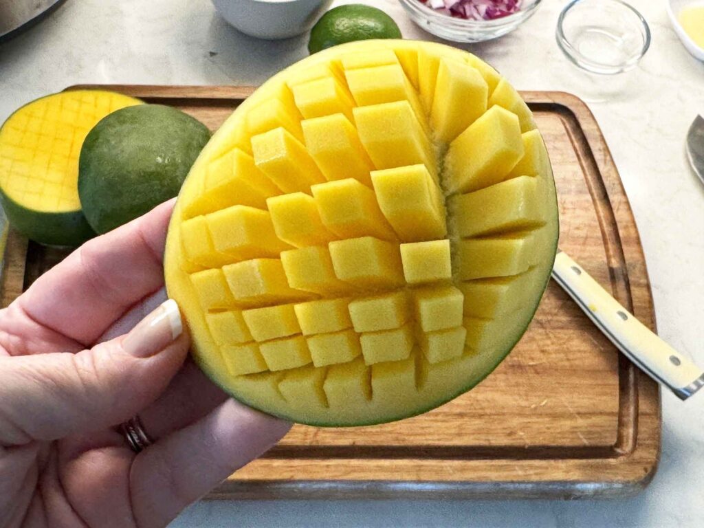 A hand holding a scored mango half cut in a crosshatch pattern over a cutting board
