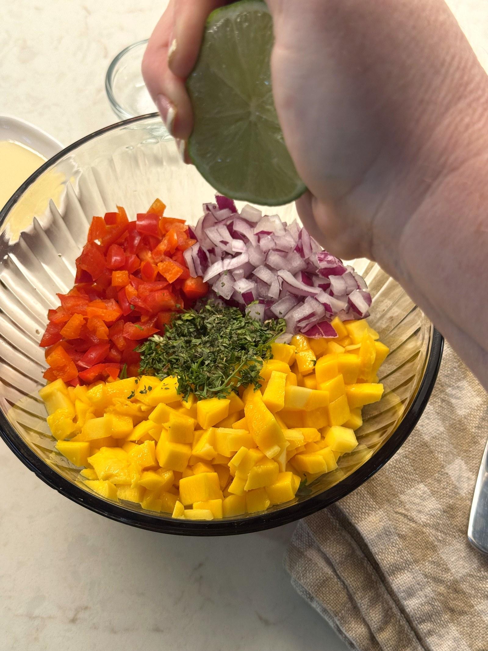 A lime being squeezed over a bowl of diced mango, bell pepper, red onion, and cilantro