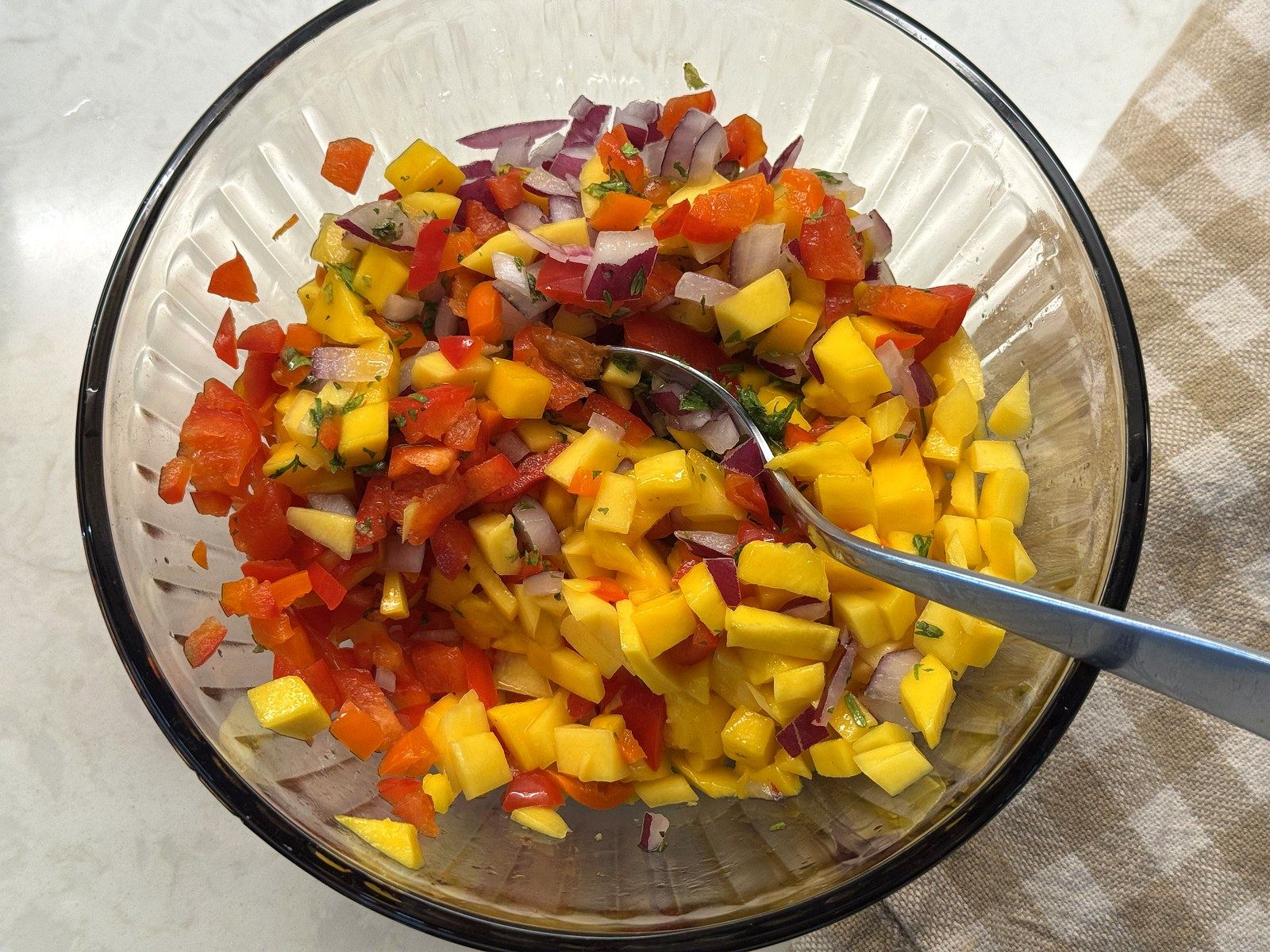 A spoon stirring together finished mango salsa in a glass bowl