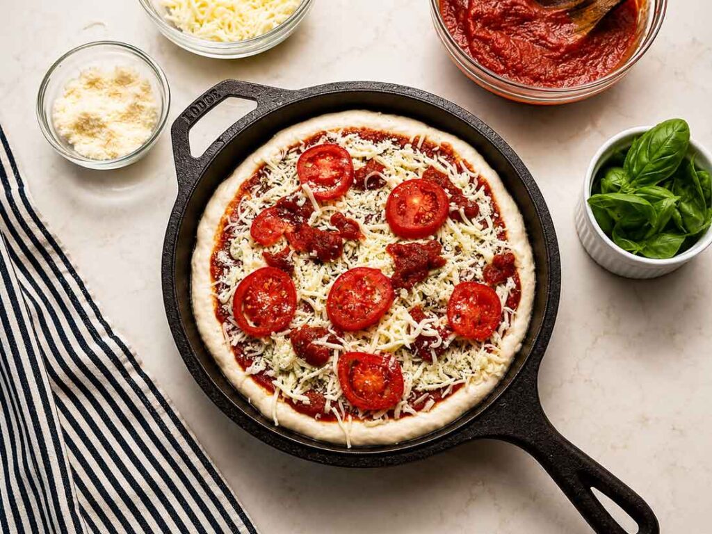 Homemade pizza sauce spread over pizza dough in a cast iron skillet and topped with shredded cheese and sliced tomatoes, with the bowl of sauce alongside