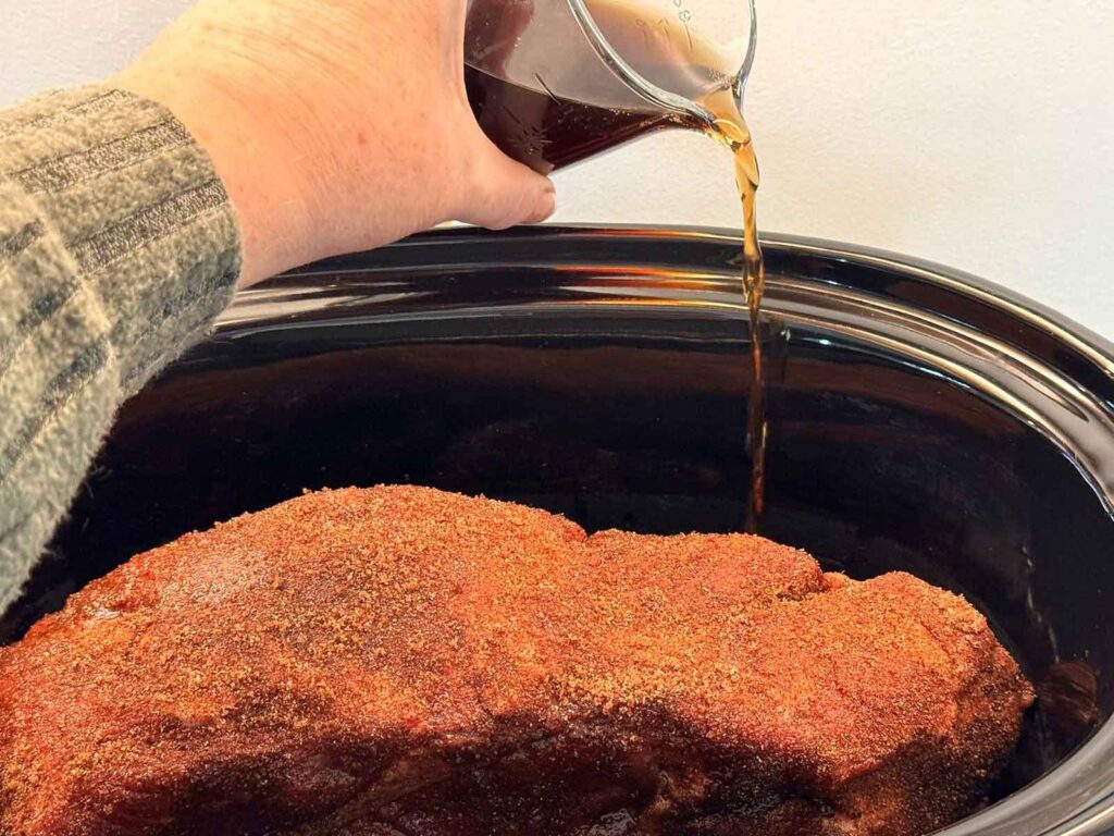 Dr Pepper being poured down the side of a slow cooker next to a seasoned pork shoulder, not over the top of the rub