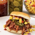 A pulled pork slider on a soft roll piled with saucy shredded pork, bread and butter pickles, and mango salsa, on a plate