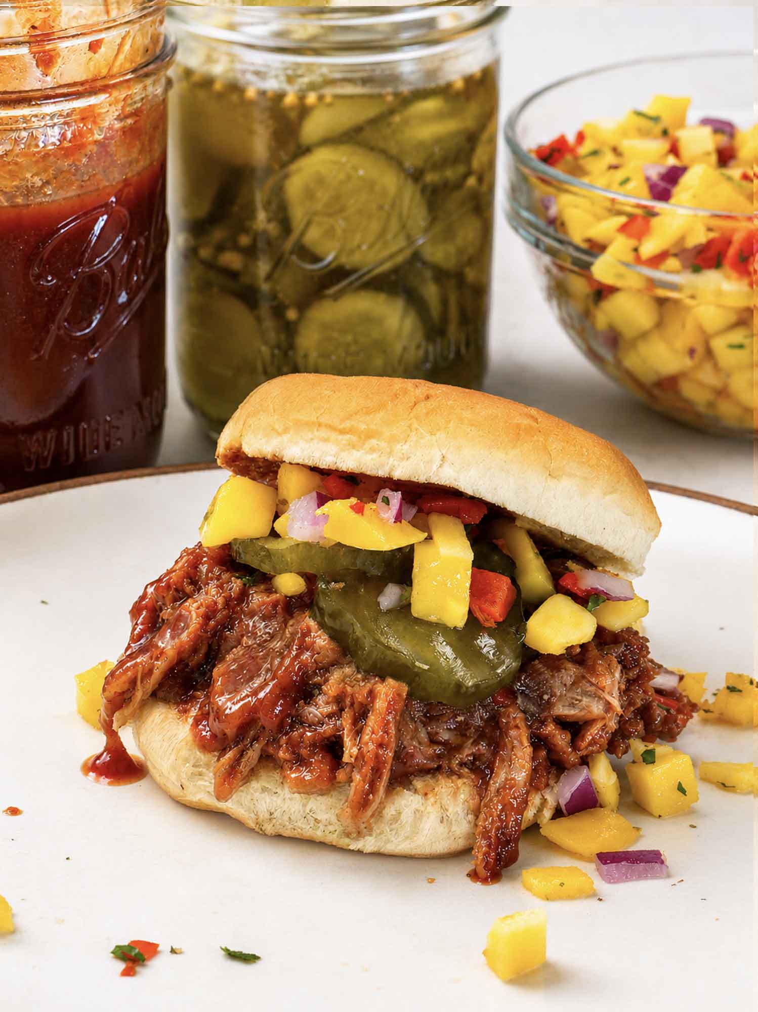 A pulled pork slider on a soft roll piled with saucy shredded pork, bread and butter pickles, and mango salsa, on a plate