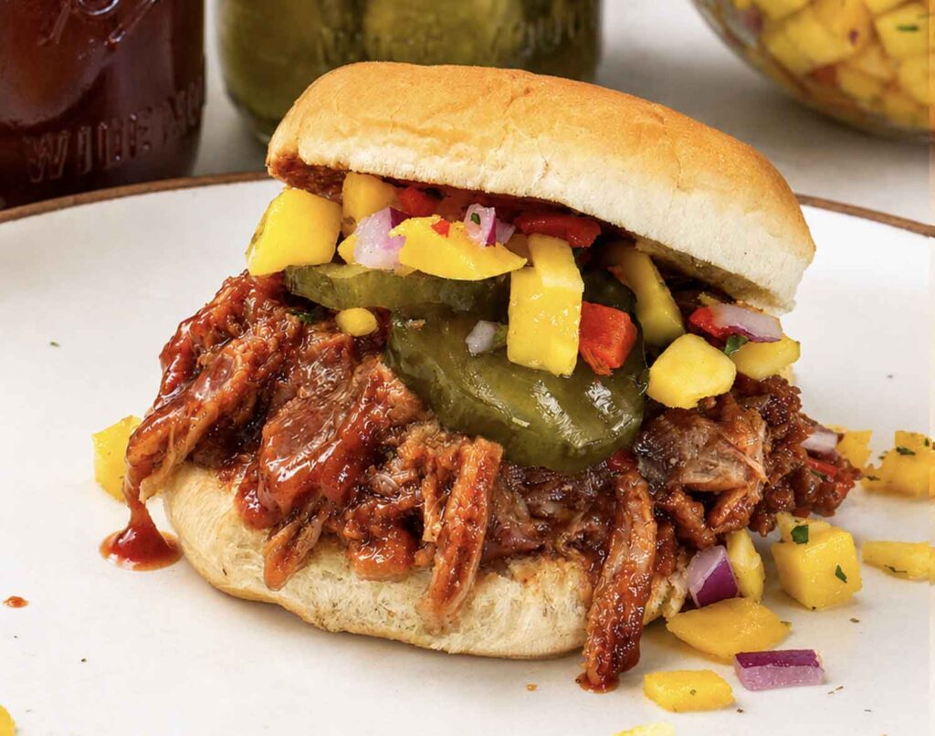 Closeup of a pulled pork slider on a soft roll piled with saucy shredded pork, bread and butter pickles, and mango salsa, on a plate