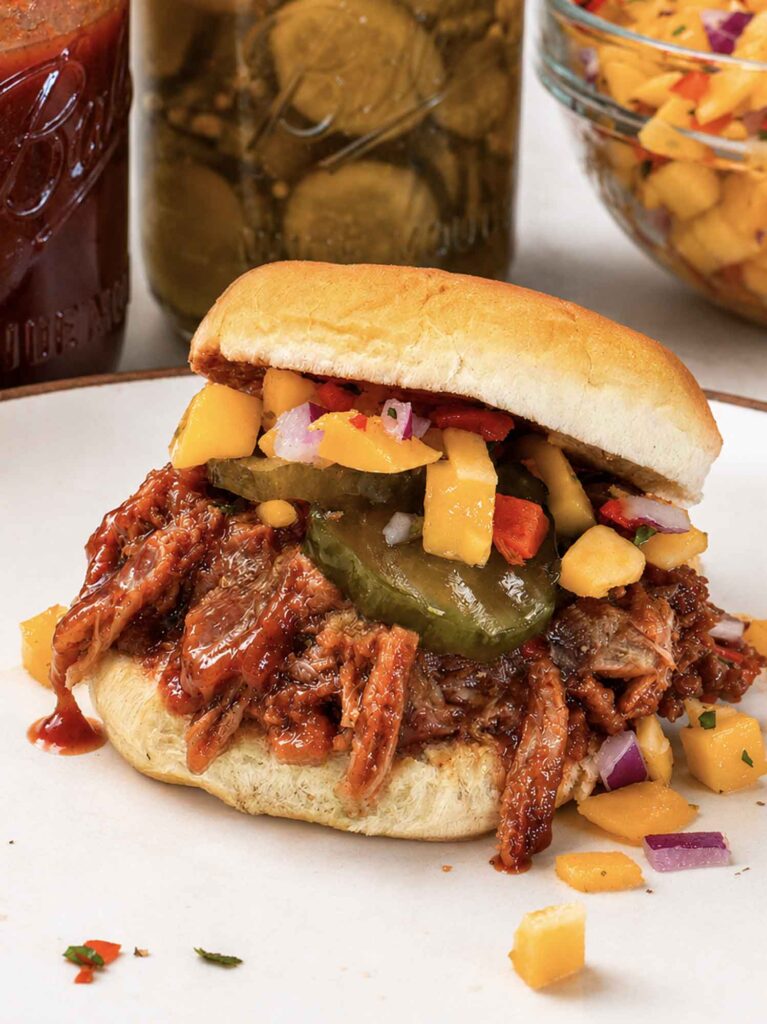 A pulled pork slider on a soft roll piled with saucy shredded pork, bread and butter pickles, and mango salsa, on a plate