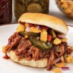 Best Pulled Pork for Sandwiches, Tacos & Sliders A pulled pork slider on a soft roll piled with saucy shredded pork, bread and butter pickles, and mango salsa, on a plate