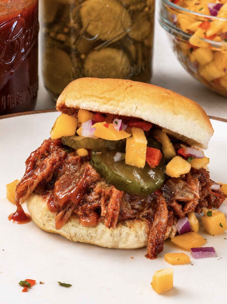 A pulled pork slider on a soft roll piled with saucy shredded pork, bread and butter pickles, and mango salsa, on a plate