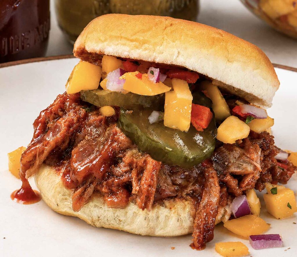 Closeup of a pulled pork slider on a soft roll piled with saucy shredded pork, bread and butter pickles, and mango salsa, on a plate