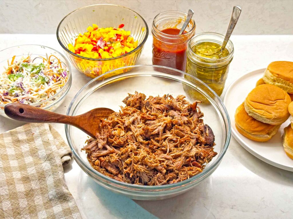 A spread of pulled pork sliders with bowls of broccoli slaw, bread and butter pickles, and mango salsa set out for serving