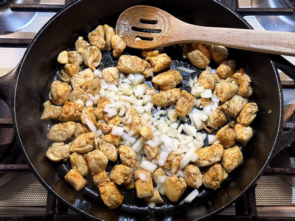 Diced onion added to the browned chicken in the skillet with a wooden spoon.