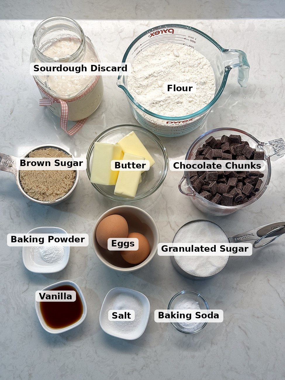 Labeled ingredients for sourdough chocolate chip cookies: flour, chocolate chunks, granulated sugar, baking soda, butter, eggs, salt, sourdough discard, brown sugar, baking powder, and vanilla.