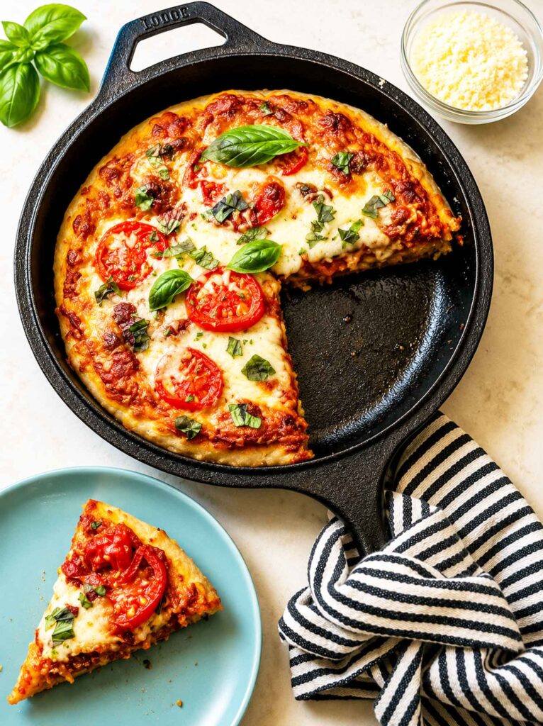 Slice of sourdough pizza on a blue plate beside a cast-iron skillet with a deep dish pizza topped with mozzarella, tomatoes, and fresh basil