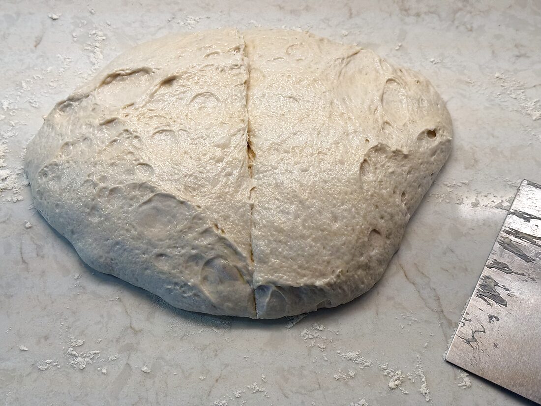 Bulk-risen sourdough pizza dough turned onto the counter and divided into two pieces with a bench scraper