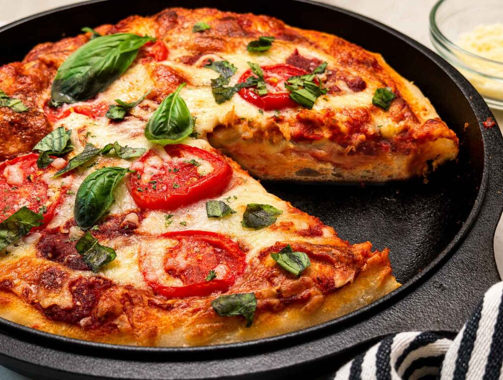 Sourdough pizza dough with a slice taken out baked into a deep dish cast iron pizza topped with mozzarella, tomatoes, and fresh basil