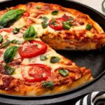 Sourdough pizza dough with a slice taken out baked into a deep dish cast iron pizza topped with mozzarella, tomatoes, and fresh basil