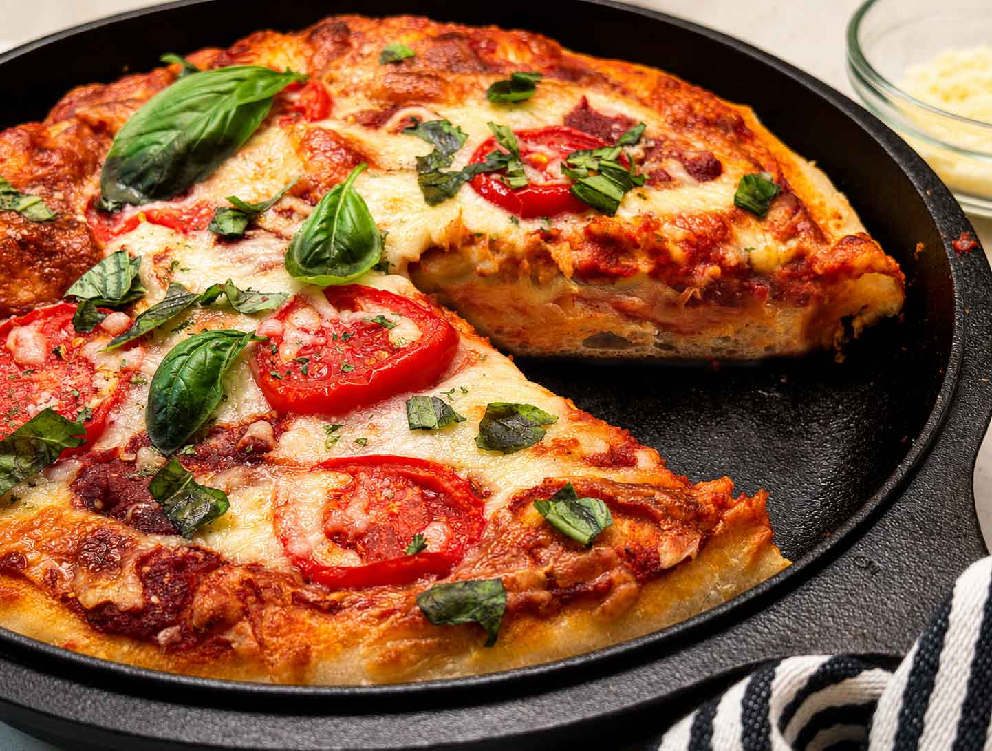 Sourdough pizza dough with a slice taken out baked into a deep dish cast iron pizza topped with mozzarella, tomatoes, and fresh basil
