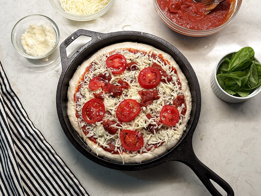 Shaped sourdough pizza crust in a cast iron skillet topped with sauce, mozzarella, and sliced tomatoes, ready to bake