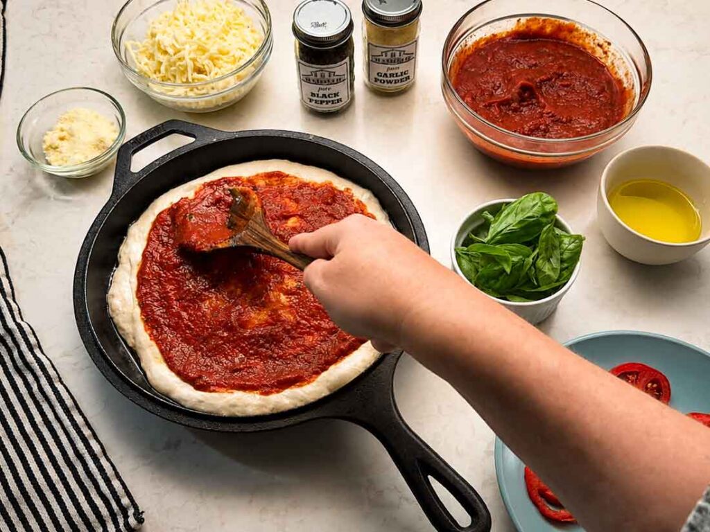 Spooning homemade pizza sauce on a sourdough pizza crust before baking
