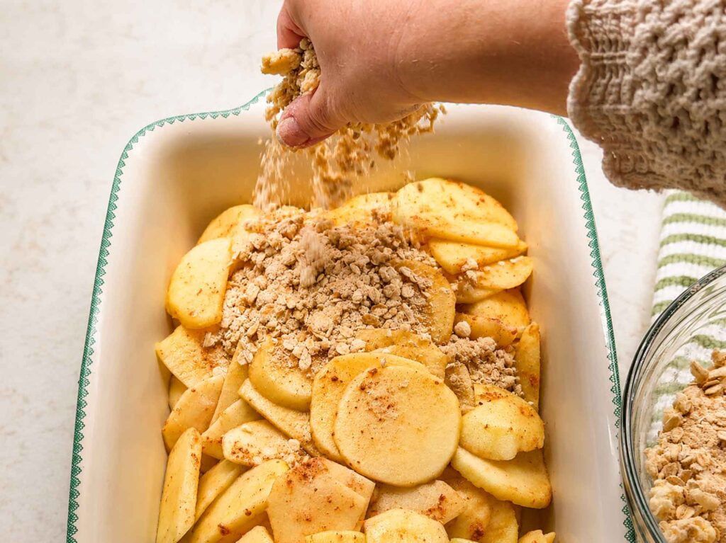 Sprinkling topping over sliced apples to make apple crisp