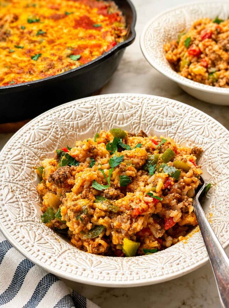 A serving of stuffed pepper casserole in a white bowl with a spoon, the skillet in the background.