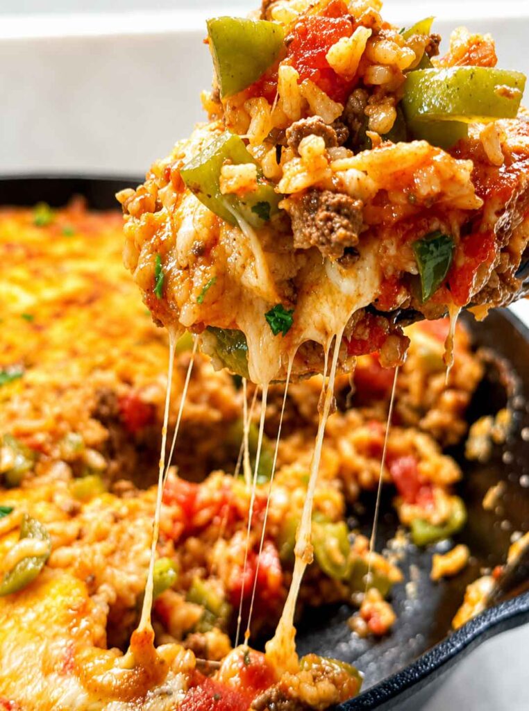 A wooden spoon lifting a scoop of stuffed pepper casserole from the skillet with melty cheese stretching.