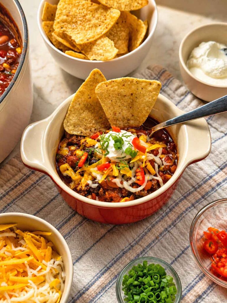 Bowl of taco soup surrounded by bowls of tortilla chips, shredded cheese, sour cream, cilantro, and red pepper