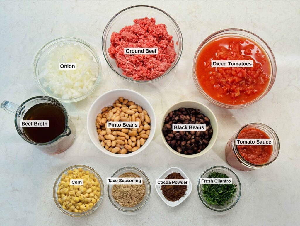 Labeled taco soup ingredients: ground beef, onion, diced tomatoes, corn, pinto beans, black beans, tomato sauce, beef broth, taco seasoning, cocoa powder, and fresh cilantro