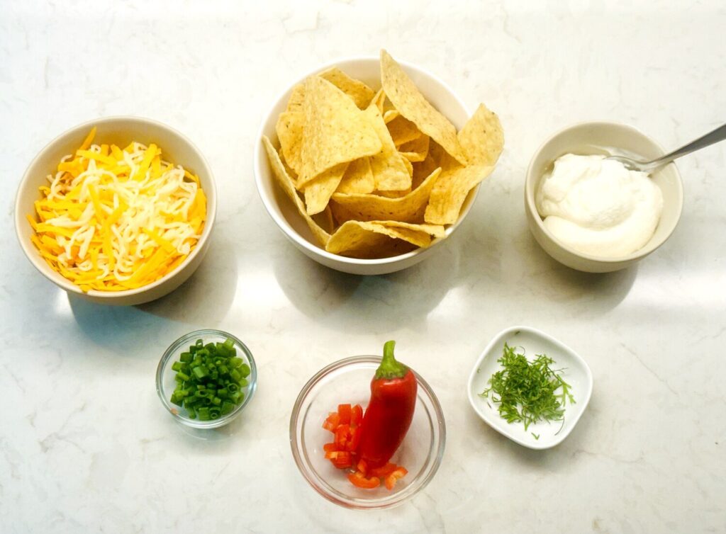 Taco soup toppings in bowls: shredded cheese, tortilla chips, sour cream, green onion, red pepper, and cilantro