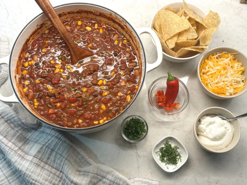 Pot of taco soup surrounded by bowls of tortilla chips, shredded cheese, sour cream, cilantro, and red pepper