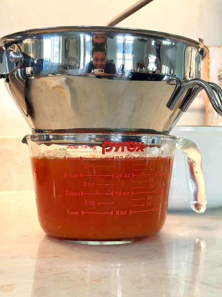 Cooked tomatoes being passed through a food mill to strain out skins and seeds into a bowl of sauce