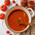 Overhead view of a pot of homemade tomato sauce with a wooden spoon, surrounded by fresh red tomatoes and two filled canning jars on a white quartz counter